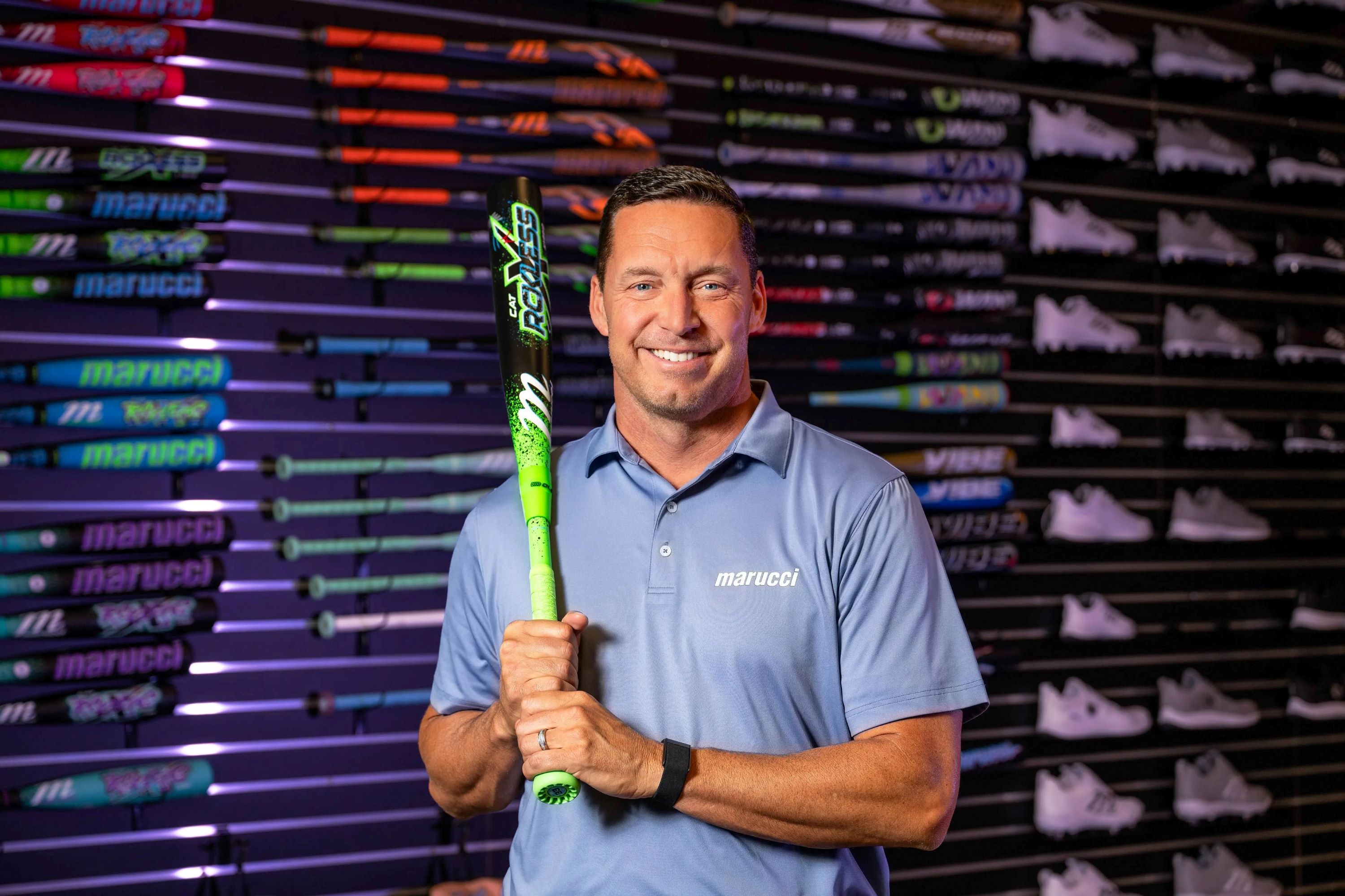 Kurt Ainsworth is holding baseball bat in front of a shelf containing bats and athletic shoes.