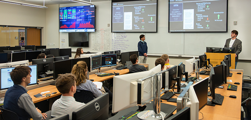 Students sit at computers in a financial lab and listen to a presentation on stocks from other students who stand under screens at the front of the room.  